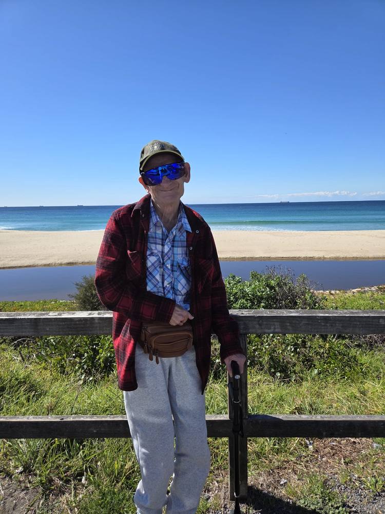 A smiling man standing by a fence overlooking the beach on a sunny day