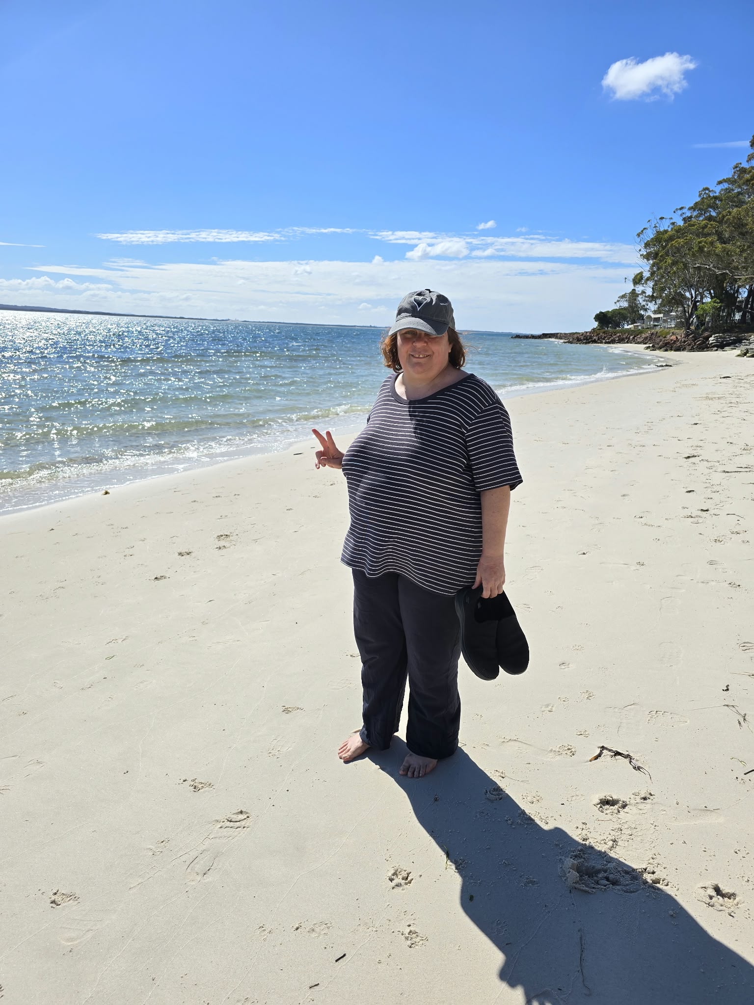 A smiling woman in a cap giving a peace sign on a sunny beach